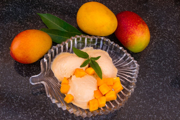 Mango ice cream with fresh mango slices with leaves mint on a glass plate. Homemade natural yellow sorbet in bowl on black granite. Vegetarian food. For gourmet. Selective focus.