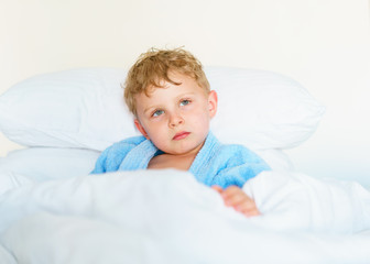 Little cute boy in blue pajamas with wet hair after bathing is lying on a huge snow-white pillow, wrapped in a blanket. He thoughtfully looks aside and positively grimaces.