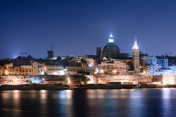 Malta's capital city Valletta, at night, from across the port in Sliema, with churches and buildings illuminated.