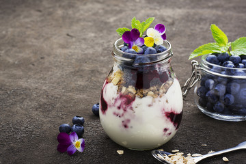 Healthy breakfast in a glass jar: yogurt, berry puree, whole grain cereal cereal, edible flowers, blueberries on a dark background. The concept of proper nutrition.