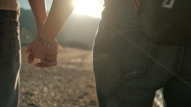 Two people in jeans walk along the rocky seashore to hold hands. At sunset, a loving couple walks along the embankment on a warm evening. Behind them they carry light black backpacks. The sun sets