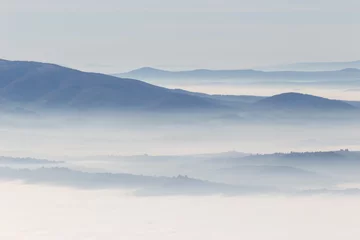 Fotobehang A view from above of a valley filled by a sea of fog, with various layers of emerging hills and mountains with different shades of blue © Massimo