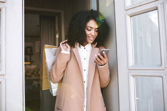 Young African American girl with dark curly hair standing with shopping bags and phone in her hands. Pretty smiling girl looking into cell phone leaning on door. Portrait of beautiful lady in coat