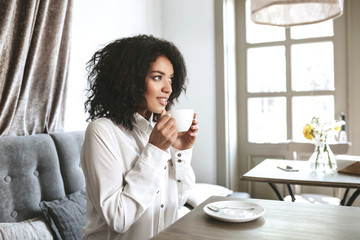 Beautiful African American girl sitting in restaurant with cup in hands. Young pretty lady in white shirt drinking coffee in cafe.Portrait of girl with dark curly hair that thoughtfully looking aside