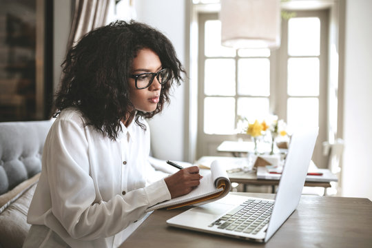 Young African American Girl Writing Notes In Restaurant.Nice Girl With Dark Curly Hair Sitting In Cafe With Laptop And Notebook.Portrait Of Lady In Glasses Thoughtfully Looking In Laptop With Notebook