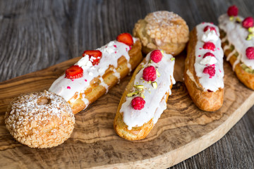 Delicious eclairs with berries and tasty shu cakes on a wooden olive board 