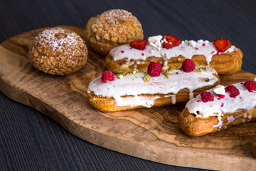 Delicious eclairs with berries and tasty shu cakes on a wooden olive board 