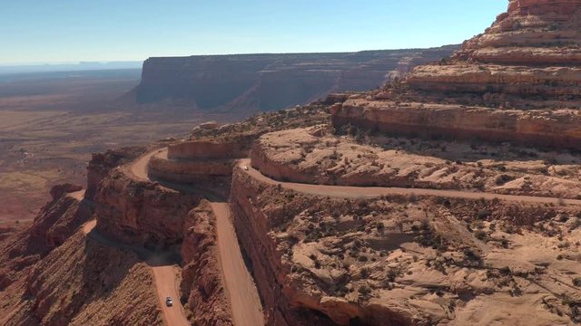 AERIAL Cars driving on sharp bend hairpin road winding up a mesa mountain cliff in red rock desert. People traveling on staggering dirt switchback Moki Dugway road carved into the face of desert cliff