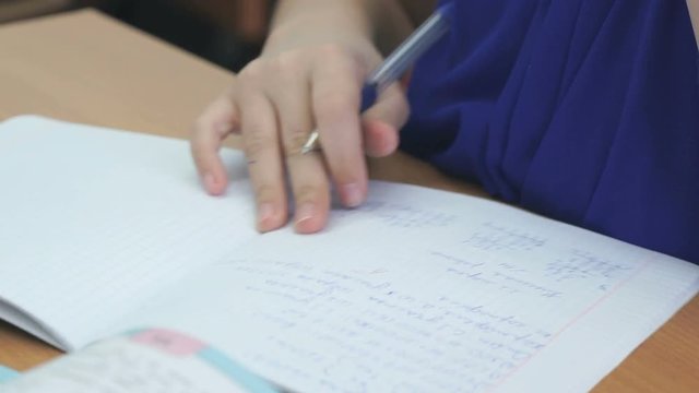 The schoolgirl sits at a school desk and writes in a notebook with a ballpoint pen. Close-up
