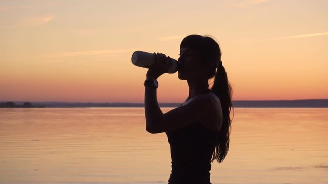 Young slender woman is drinking water from a bottle on the lake at sunset