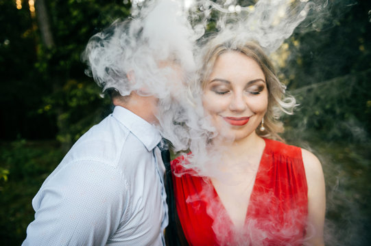 Smoking businessman kissing beautiful young blonde girl with innocent kind face, piercing, eyes closed, red lips, dress with decolette. Clouds of thick smoke. Electronic cigarette. Odd lovers feelings