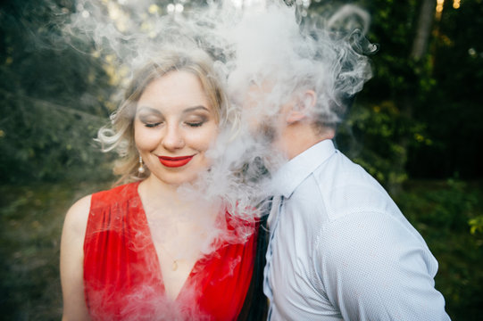 Smoking businessman kissing beautiful young blonde girl with innocent kind face, piercing, eyes closed, red lips, dress with decolette. Clouds of thick smoke. Electronic cigarette. Odd lovers feelings