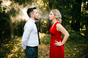 Businessman and beautiful blonde woman with coiffure in red expensive dress with earrings standing in forest with electronic cigarrette and blow out smoke in front each other. Smoking addiction. Park.