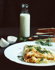 Tasty omelette with greens on a white plate with a glass of milk, bread and chicken eggs on a dark wooden background