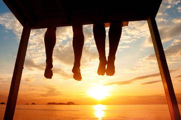 happiness concept, silhouette of feet of couple sitting on the pier at sunset beach - Powered by Adobe