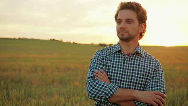 Close up portrait of handsome man standing in green wheat field on sunset and looking at the camera.