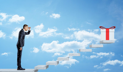 A businessman on sky background looking up at a grey stairway to see a white giftbox with a red bow.