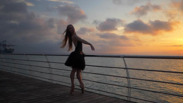 Beautiful scene of a dancing ballerina in black ballet tutu and pointe on embankment above ocean or sea beach at sunrise or sunset. Young beautiful blonde woman with long hair practicing stretching