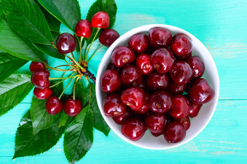 Ripe juicy red cherries in a ceramic bowl on a bright wooden background, among the leaves. Top view.