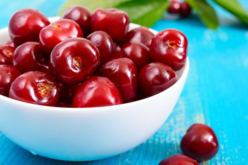 Ripe red cherry berries in a white ceramic bowl on a blue wooden background.