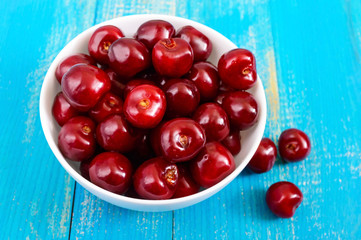 Ripe red cherry berries in a white ceramic bowl on a blue wooden background.