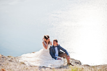 Stunning young wedding couple sitting on the edge of the cliff with a beautiful scenery on the background.