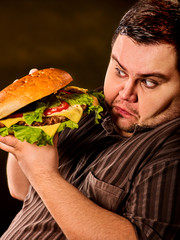 Man eating fast food hamberger. Fat person with brutal look made great huge hamburger and admires him, intending to eat it. Junk meal leads to obesity. Person regularly overeats concept .