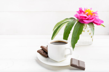 White cup of coffee with flower in vase and chocolate on whte wooden background