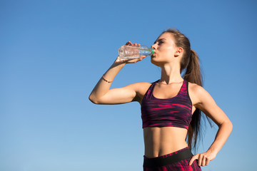 Young happy woman drinking water from bottle. Isolated sportswoman in sportswear holding drink outdoors at blue sky background. She quenches thirst.