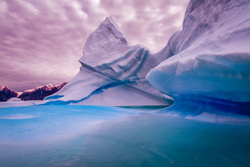 Obrazy na Szkle  Iceberg streaked with patterns of dark blue ice with frozen blue ice in the foreground and a glacier with pink sky behind.