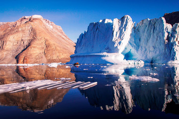 Blue and chocolate iceberg with layered mountain behind reflected in the water in front with ribbed ice and an iceberg. © Di
