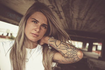 Portrait of a young beautiful blonde in a white T-shirt and with tattoos, looks at the camera,...