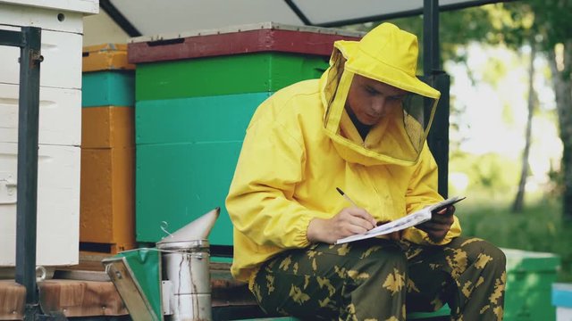 Young beekeeper man write in notepad checking harvest while sitting near beehives