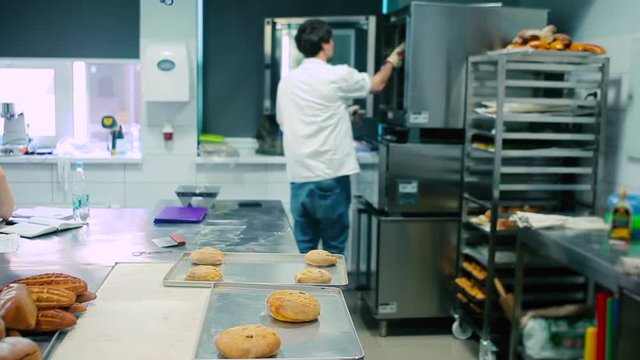 The cook open the stove. The man take away the bread from the stove. The cook put the bread in the trays at the table. The cook take one bread into the hand.