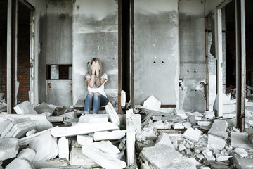 Fototapeta na wymiar A lonely girl in a white T-shirt and jeans sits on the ruins of a ruined house. Sad expression, tragic atmosphere