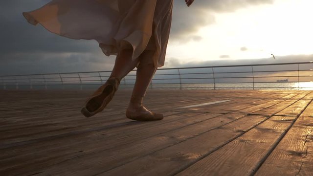 Young ballerina in beige silk dress running in pointe shoes on wooden embankment to sea or ocean. Sunrise background. Slow motion.