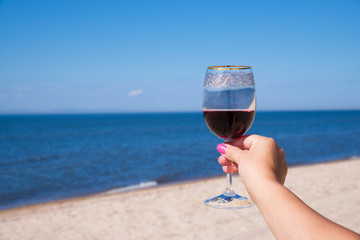 A glass of red wine in a beautiful female hand with pink nails. Against the blue sunny sky and the sea