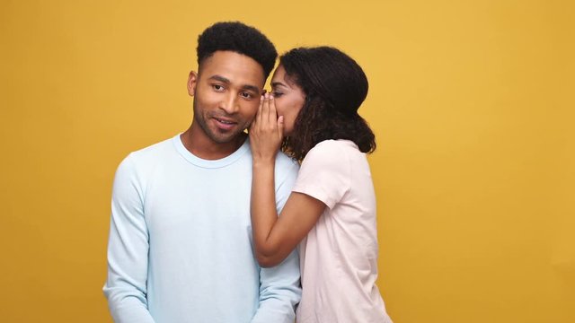 Young african woman telling a big secret to her shocked boyfriend isolated over yellow background