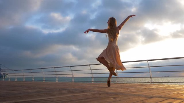 Dancing ballerina in beige silk dress and pointe on embankment above ocean or sea beach at sunrise or sunset. Young beautiful blonde woman with long hair practicing stretching and classic exercises