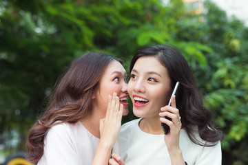Surpried young woman whispering at a cheerful friend while on call