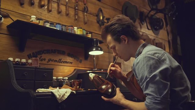 Young bootmaker smearing special paint liquid with a brush on a shoe while sitting at his table at the workshop
