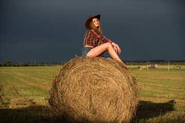 Naklejka na ściany i meble Young woman in shorts and cowboy hat sitting on a haymow