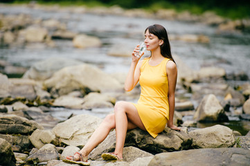 Young lonely tourist cutie girl sitting on stone on shore of river in mountains in wild terrain and satisfying thirst drinking water from glass. Traveler resting at nature. Fluid balance. Heat weather
