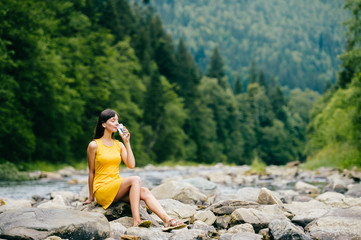 Young lonely tourist cutie girl sitting on stone on shore of river in mountains in wild terrain and satisfying thirst drinking water from glass. Traveler resting at nature. Fluid balance. Heat weather