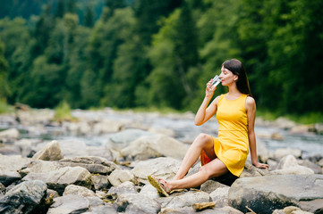 Young lonely tourist cutie girl sitting on stone on shore of river in mountains in wild terrain and satisfying thirst drinking water from glass. Traveler resting at nature. Fluid balance. Heat weather
