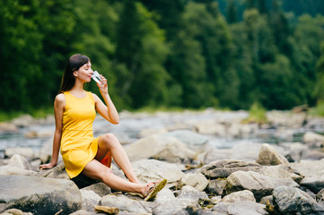 Young lonely tourist cutie girl sitting on stone on shore of river in mountains in wild terrain and satisfying thirst drinking water from glass. Traveler resting at nature. Fluid balance. Heat weather