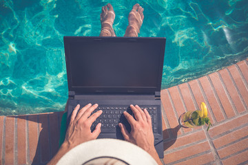Tourist sitting at the edge of swimming pool and using blank screen laptop. Summer vacation and freelancer concepts