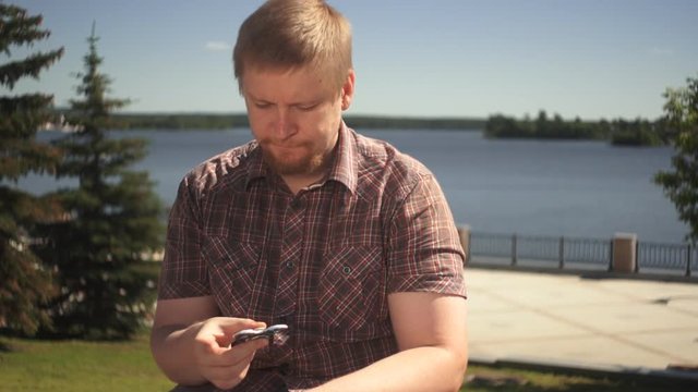 A young red-bearded man in a checkered shirt twists in the hands of a toy spiner sitting on the waterfront background of a blue pond on a warm summer day.