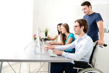 Obraz premium disabled handicapped young male worker on wheelchair with coworker in business office working on a desktop computer
