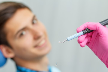 Doctor wearing pink gloves is holding ultrasonic dental scaler with young man smiling on the background